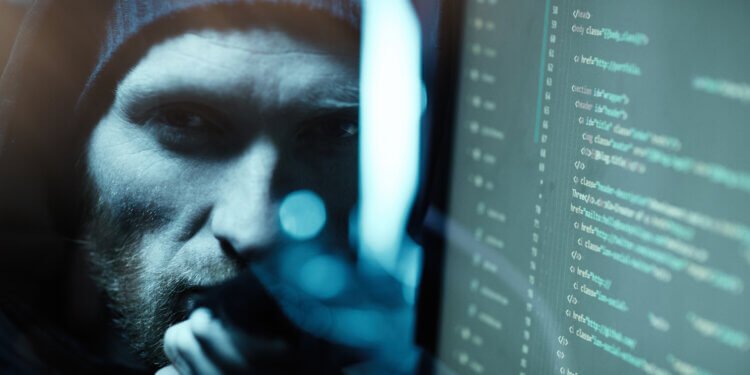 Portrait of young bearded computer programmer looking at camera near the computer monitor with software in dark office