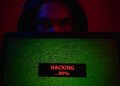 A hooded person’s face, lit in red and blue, peers over a laptop showing dense green code. A status box reads “HACKING…99%.” A dark, semi‑transparent panel overlays the image with the headline “Salesforce Connected‑App Data Heist — What Happened & How to Protect Yourself” and “ScamShieldDigest.com · September 2025.”