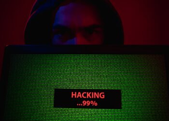 A hooded person’s face, lit in red and blue, peers over a laptop showing dense green code. A status box reads “HACKING…99%.” A dark, semi‑transparent panel overlays the image with the headline “Salesforce Connected‑App Data Heist — What Happened & How to Protect Yourself” and “ScamShieldDigest.com · September 2025.”
