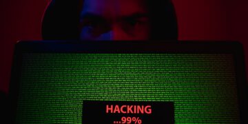 A hooded person’s face, lit in red and blue, peers over a laptop showing dense green code. A status box reads “HACKING…99%.” A dark, semi‑transparent panel overlays the image with the headline “Salesforce Connected‑App Data Heist — What Happened & How to Protect Yourself” and “ScamShieldDigest.com · September 2025.”