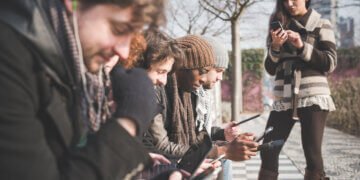 Six young adult friends using smartphones and digital tablets in park